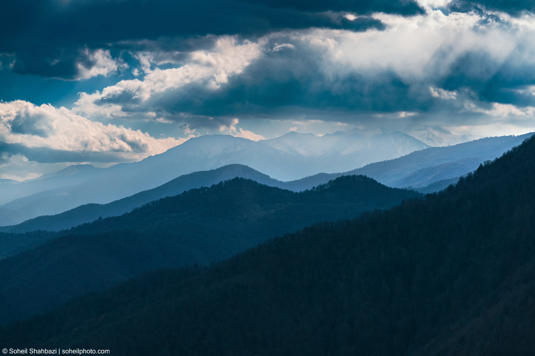 A wide landscape shot of layered blue mountain ridges receding into the distance under a dramatic sky with heavy, dark clouds and patches of light. Photographed by Soheil Shahbazi