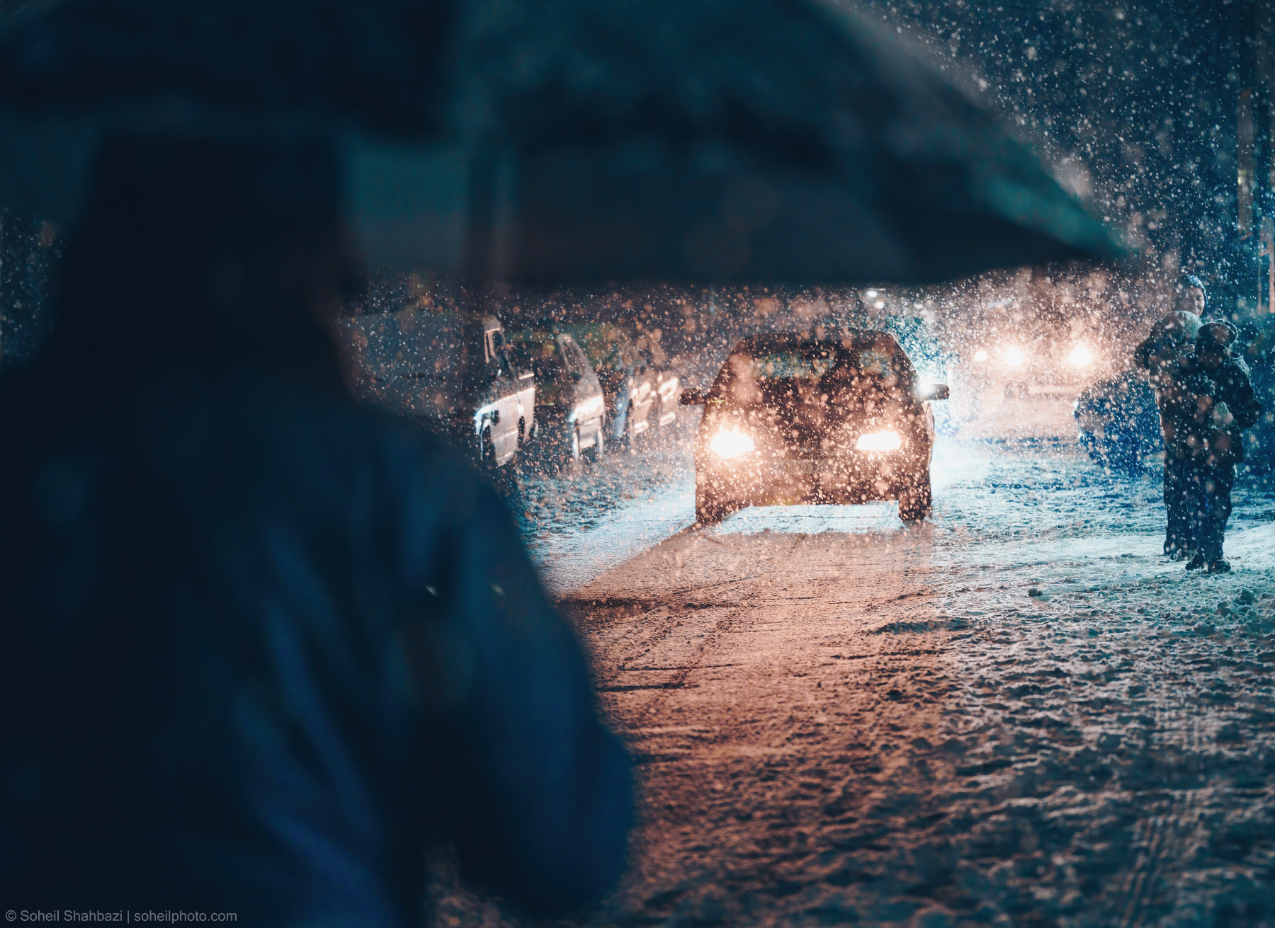 Cinematic night shot of a snowy street with cars and their headlights illuminating the falling snow photographed by Soheil Shahbazi