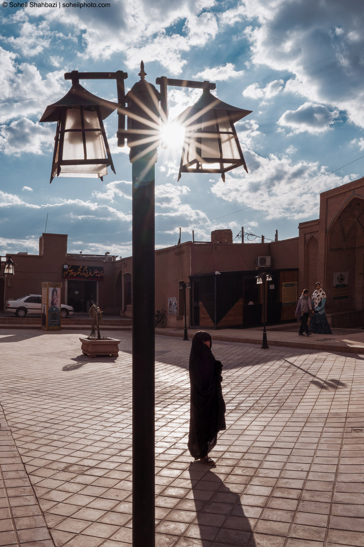 A dramatic low-angle shot of a woman in a black chador standing on a paved square, positioned behind a lamp post with the sun creating a bright starburst effect between the lanterns. Photographed by Soheil Shahbazi