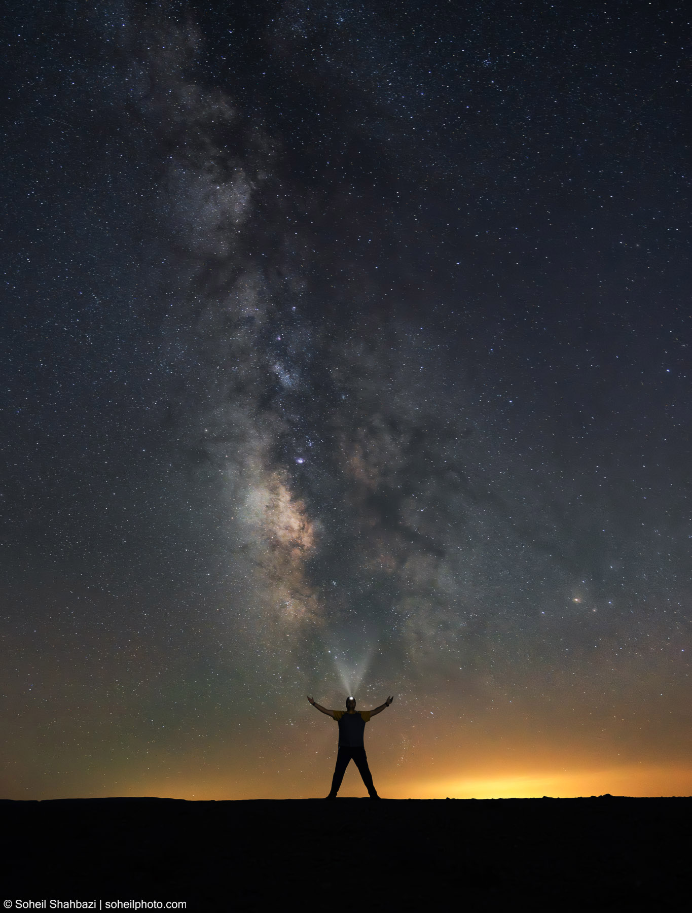 Silhouette of a man standing with open arms under a breathtaking night sky featuring the detailed Milky Way galaxy and a starry horizon - Photographed by Soheil Shahbazi