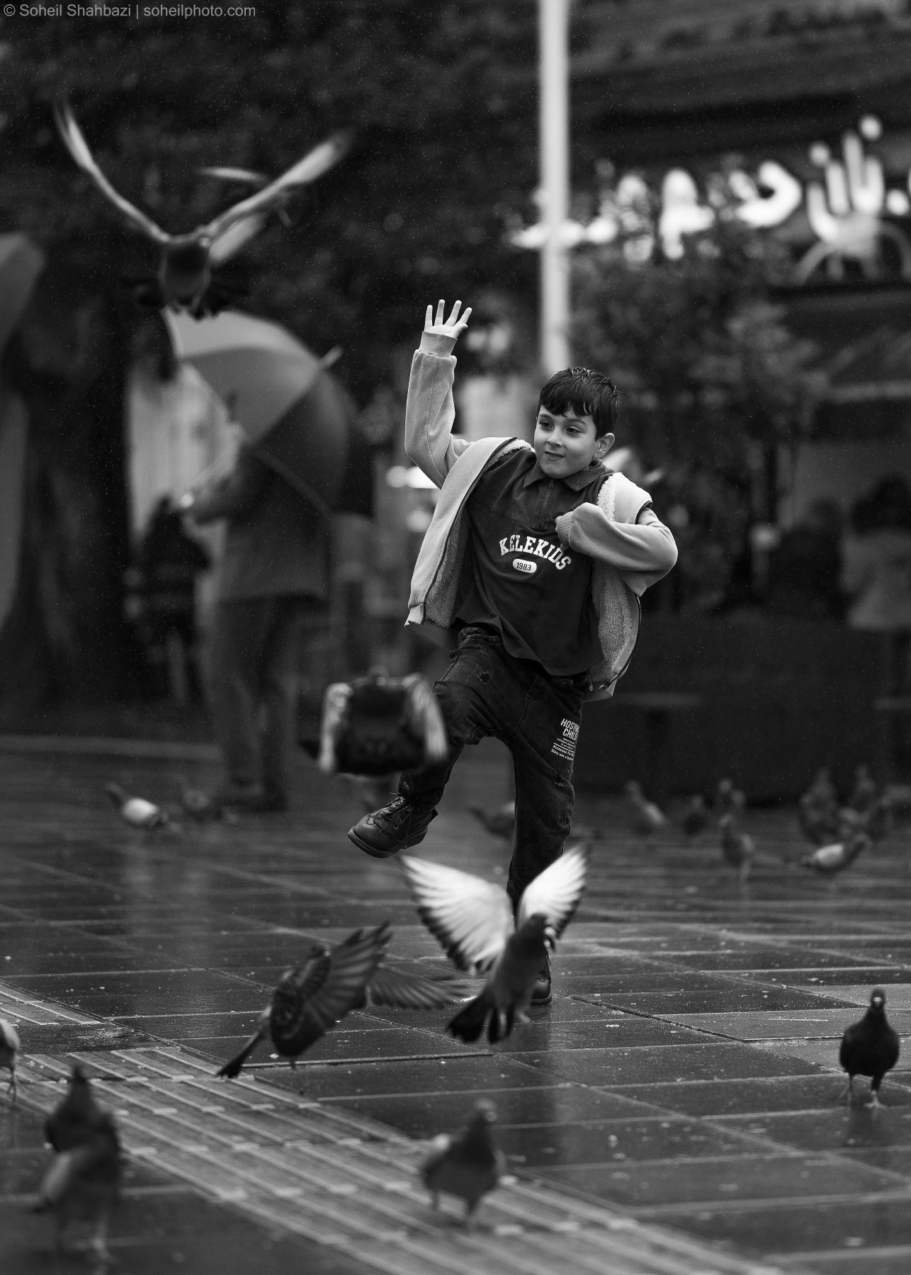 Black and white action shot of a joyful young boy playing and jumping among a flock of pigeons on a wet city pavement, photographed by Soheil Shahbazi
