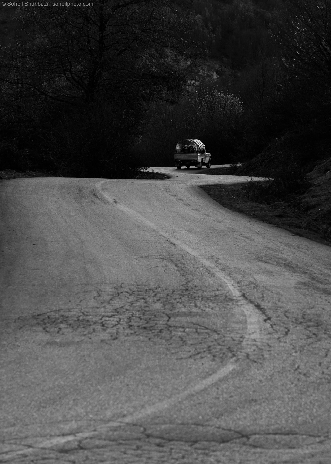 A black and white cinematic shot of a winding road leading towards a small truck in the distance, surrounded by dark trees and a moody atmosphere. Photographed by Soheil Shahbazi