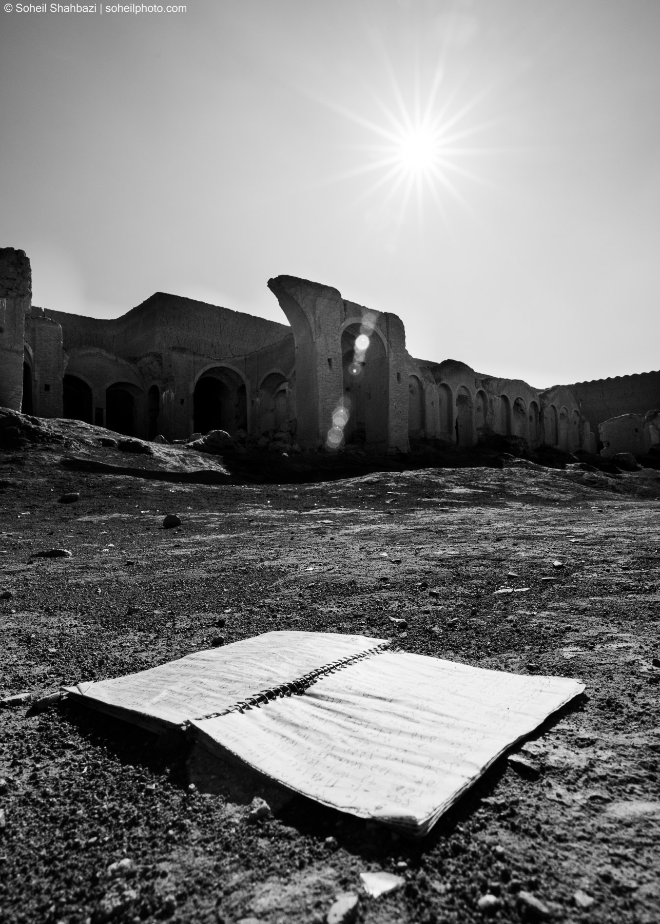 Alt text: A dramatic black and white low-angle shot of an open notebook lying on the sandy ground in front of ancient mud-brick ruins under a bright, piercing sun, photographed by Soheil Shahbazi