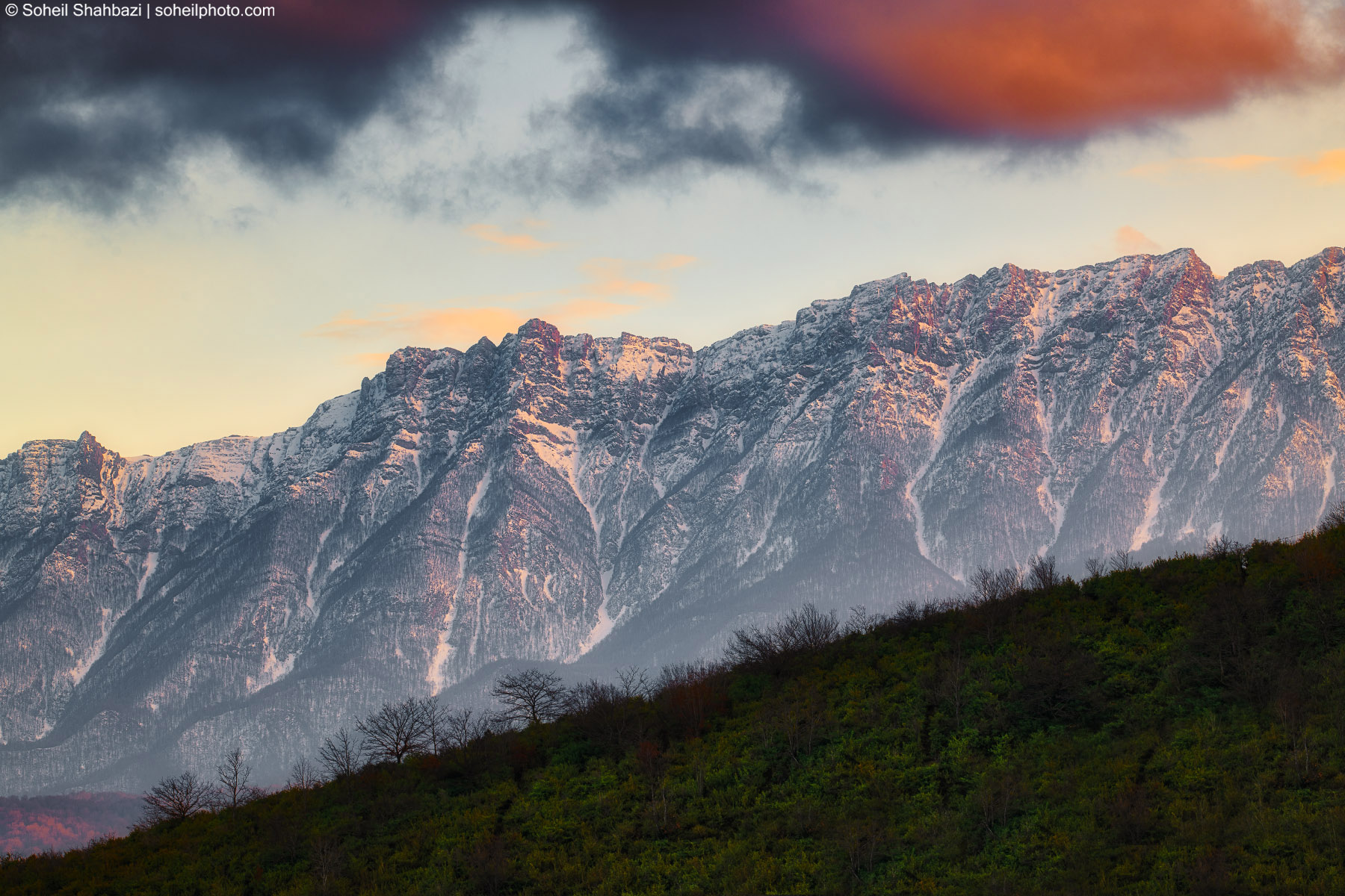 A majestic landscape shot of snow-capped mountain peaks glowing under the warm light of a sunset or sunrise, contrasting with dark dramatic clouds above and a lush green hillside in the foreground. Photographed by Soheil Shahbazi