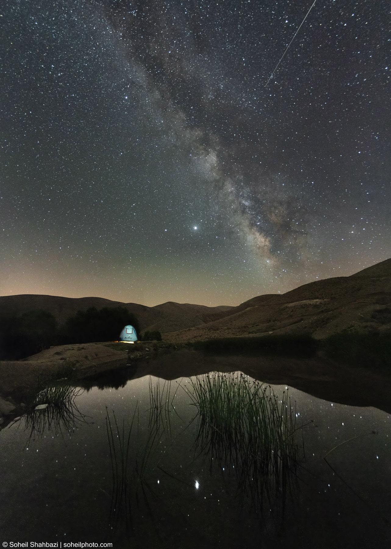 A wide-angle night landscape featuring a luminous tent reflecting in a still pond, with the majestic Milky Way galaxy stretching across a star-filled sky over rolling hills - Photographed by Soheil Shahbazi