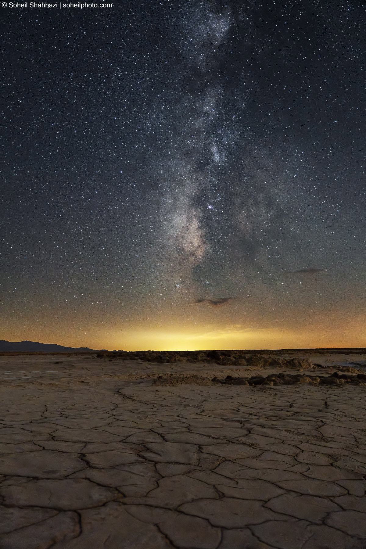 A vast night landscape of a cracked dry lake bed under a stunningly detailed Milky Way galaxy, with a golden glow on the horizon - Photographed by Soheil Shahbazi