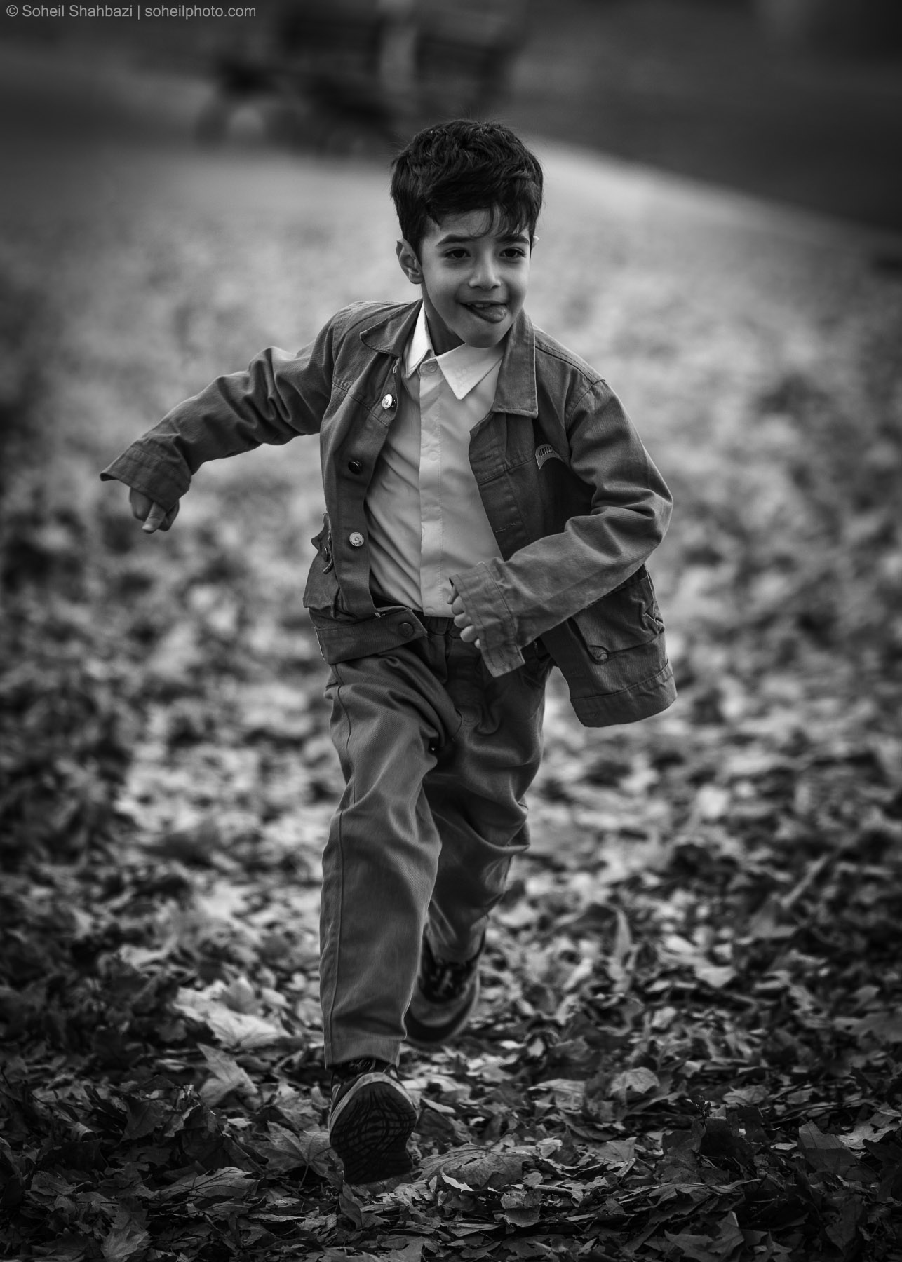 Black and white candid shot of a joyful young boy walking through fallen autumn leaves by Soheil Shahbazi