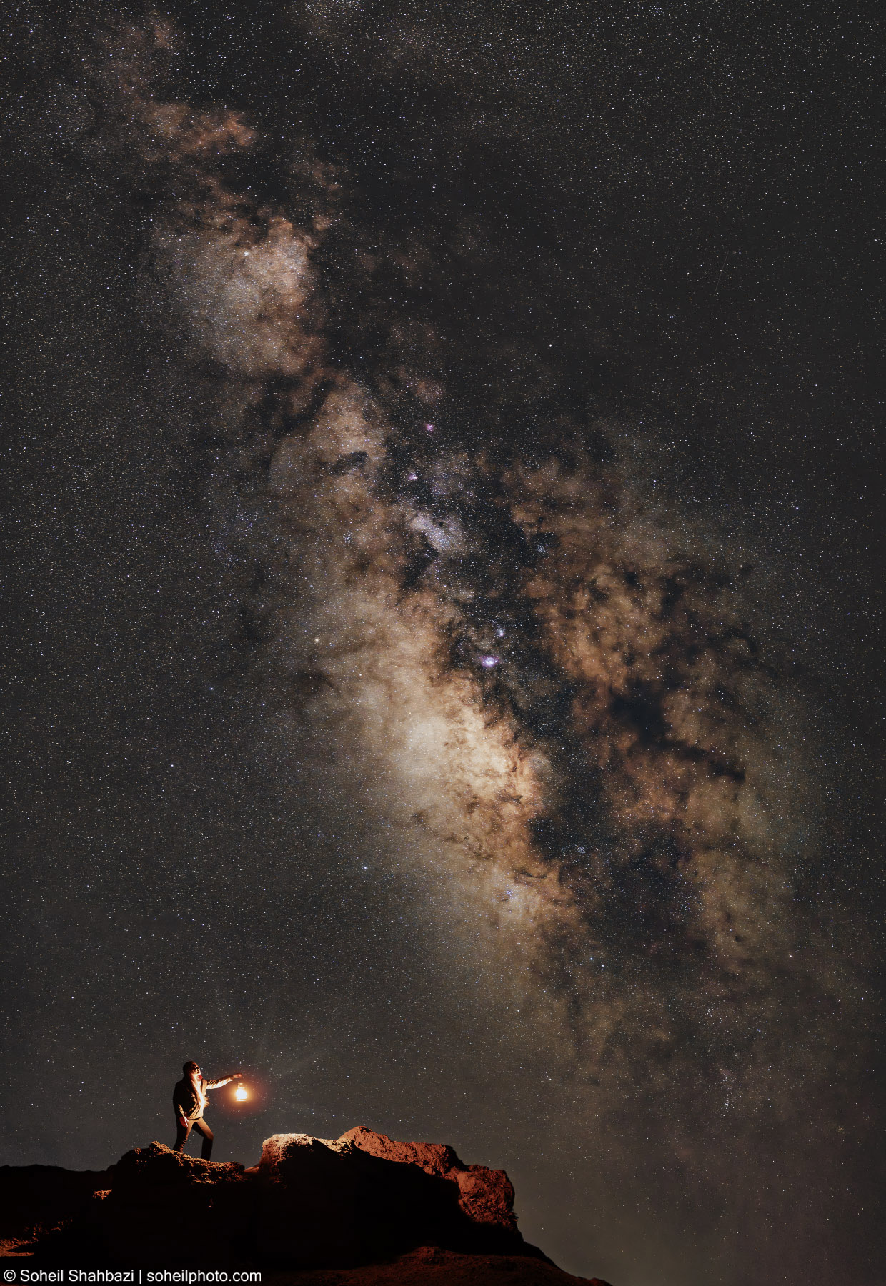 Silhouette of a person standing on a rocky peak at night, holding a light source that illuminates the ground, set against a vast and detailed view of the Milky Way galaxy - Photographed by Soheil Shahbazi