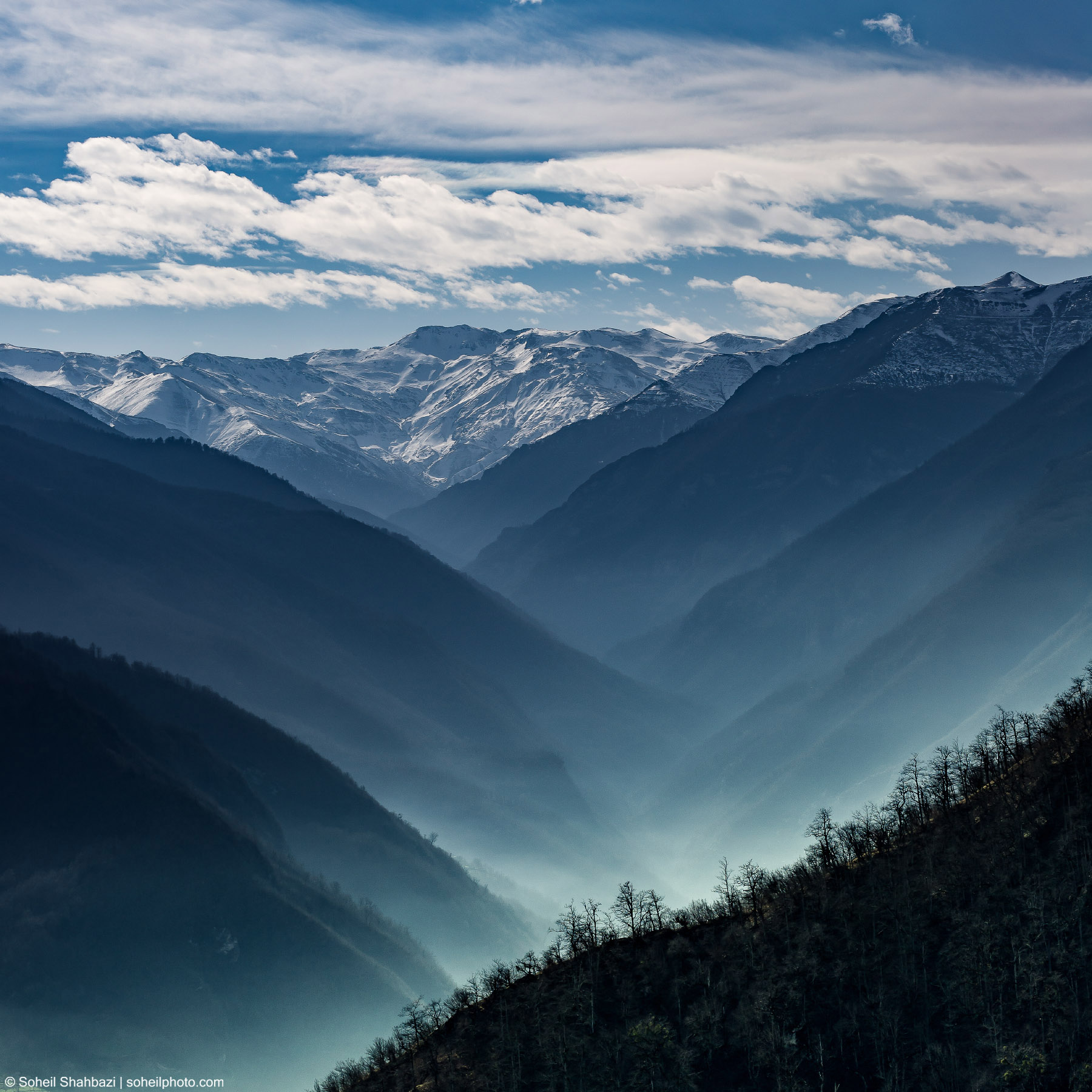 Dramatic landscape of layered blue mountains with snow-capped peaks under a bright cloudy sky, featuring a deep valley filled with soft mist - Photographed by Soheil Shahbazi