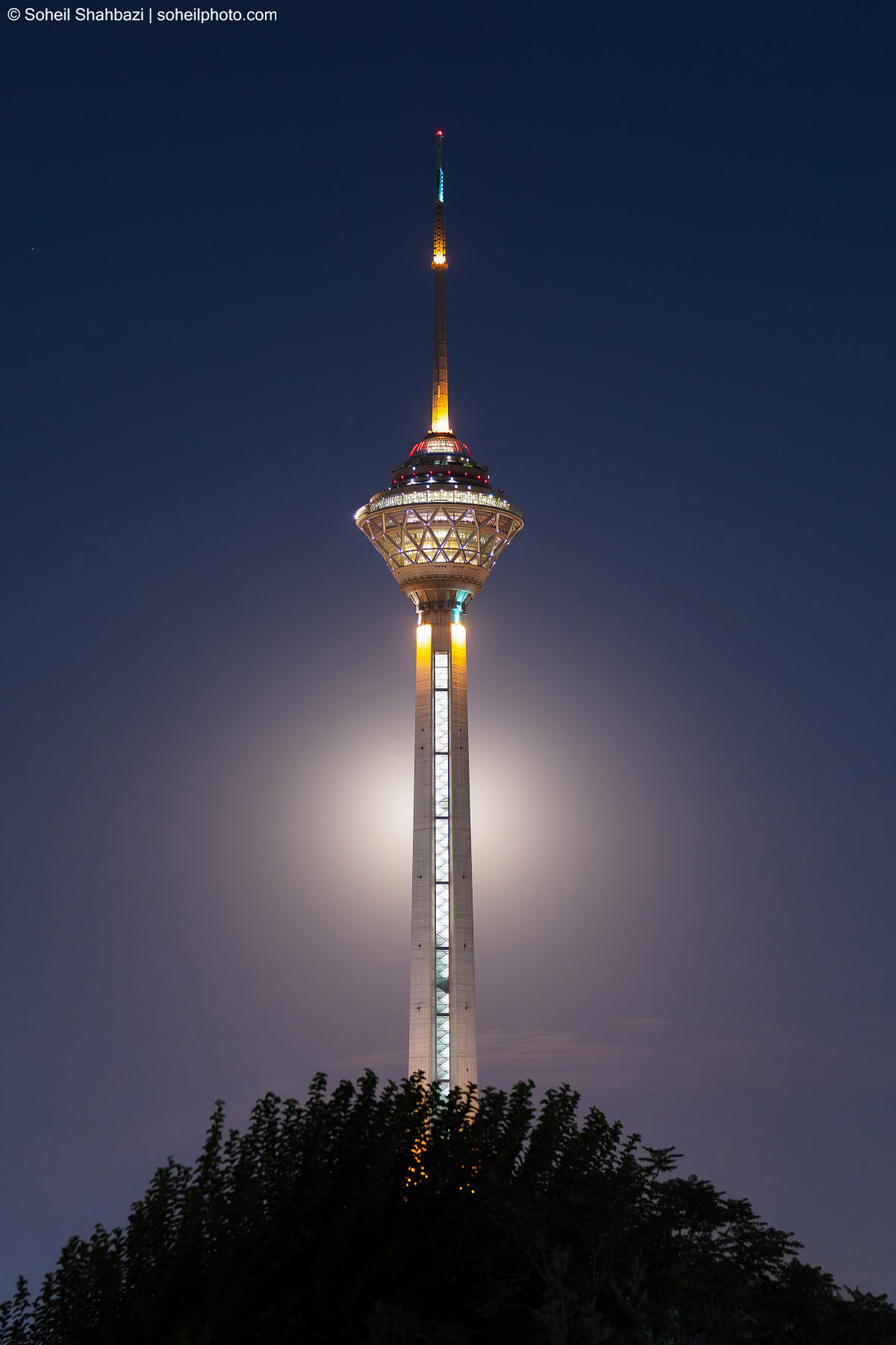 Milad Tower illuminated at night against a dark blue sky, standing tall above a silhouette of lush green trees with a bright glow behind the tower - Photographed by Soheil Shahbazi