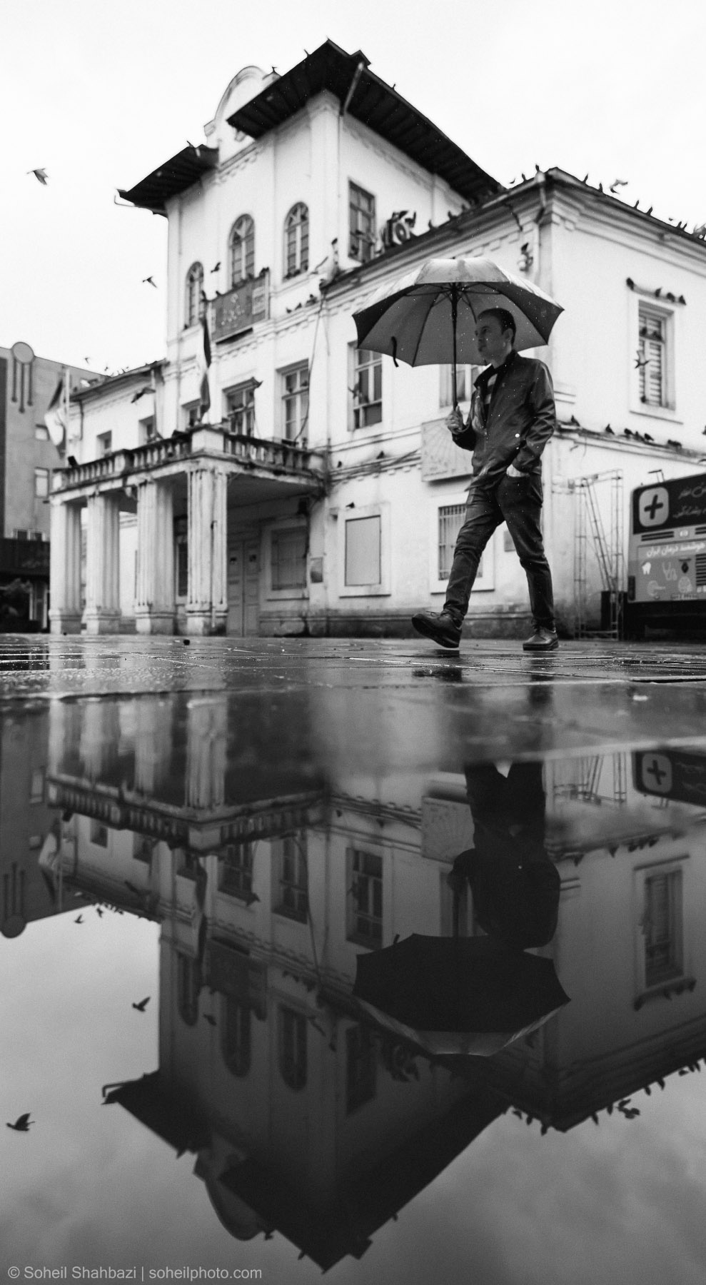 Alt text: A black and white street photograph of a man in a suit walking with an umbrella in front of a vintage building, with a perfect symmetrical reflection in a rain puddle on the ground, photographed by Soheil Shahbazi
