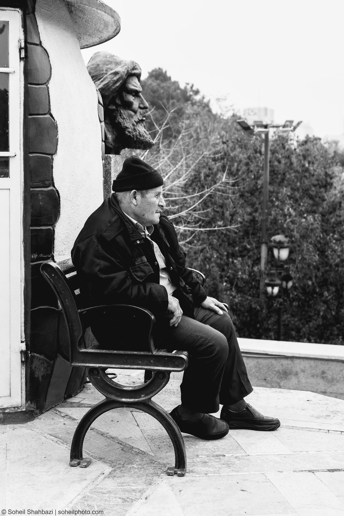 Alt text: A black and white side profile shot of an elderly man wearing a beanie and jacket sitting on a bench, with a large stone sculpture of a face positioned directly above him, creating a visual parallel, photographed by Soheil Shahbazi