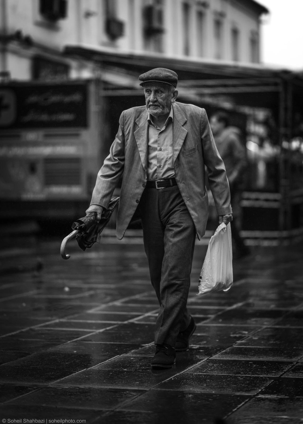 Black and white street portrait of an elderly man wearing a flat cap and a suit, walking on a wet pavement while carrying an umbrella and a plastic shopping bag, photographed by Soheil Shahbazi