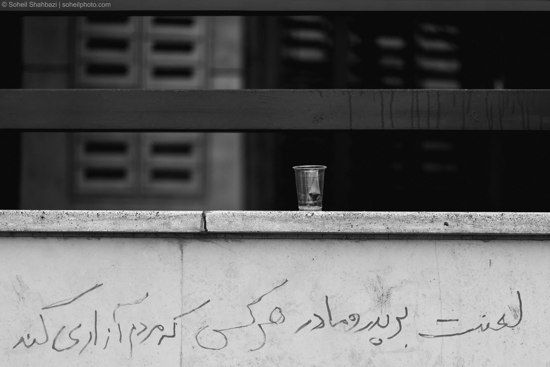 A black and white cinematic shot of a single disposable plastic cup sitting on a concrete ledge, with Persian calligraphy graffiti written on the wall below it and a blurred urban background. Photo by Soheil Shahbazi.