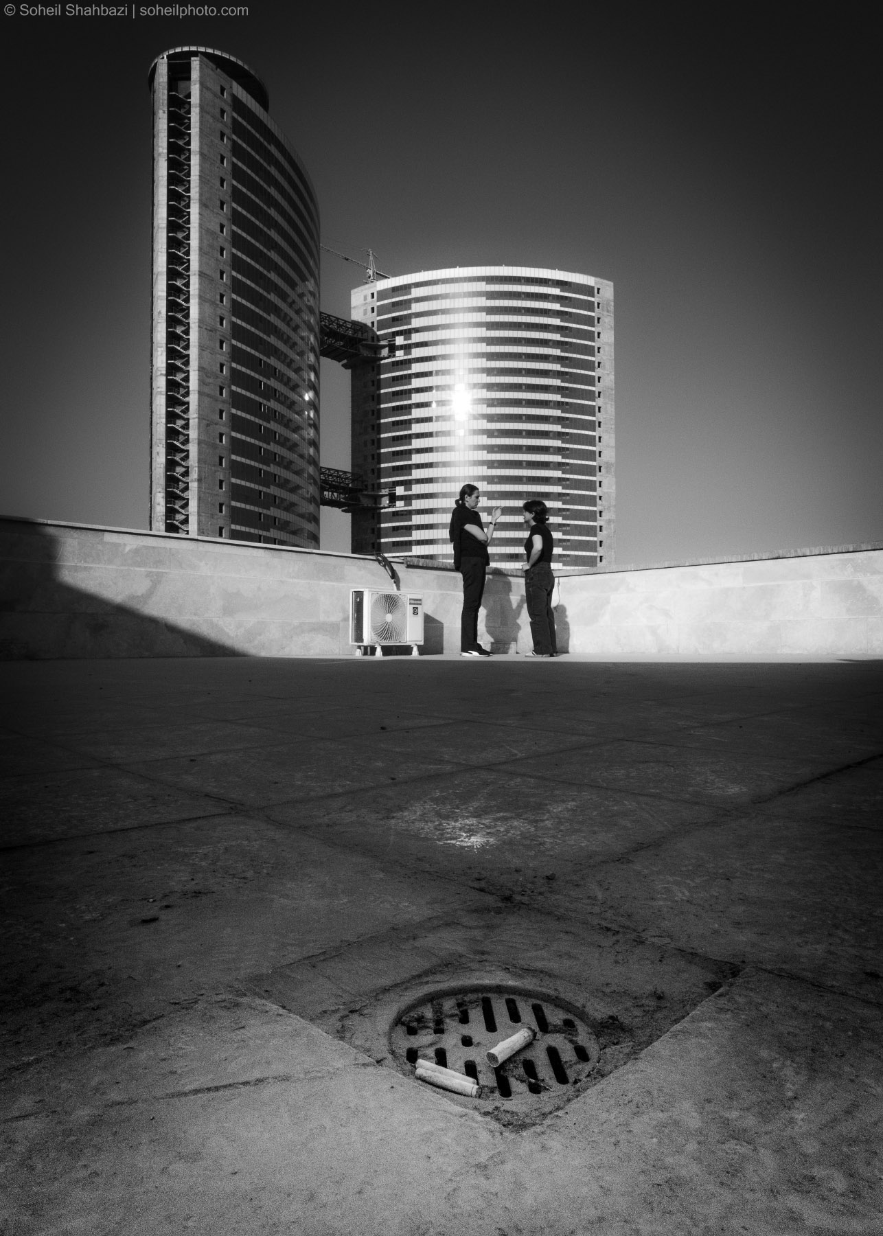 A high-contrast black and white architectural shot featuring two people standing in the distance against a backdrop of two tall, cylindrical modern buildings, with a manhole cover in the foreground. Photographed by Soheil Shahbazi