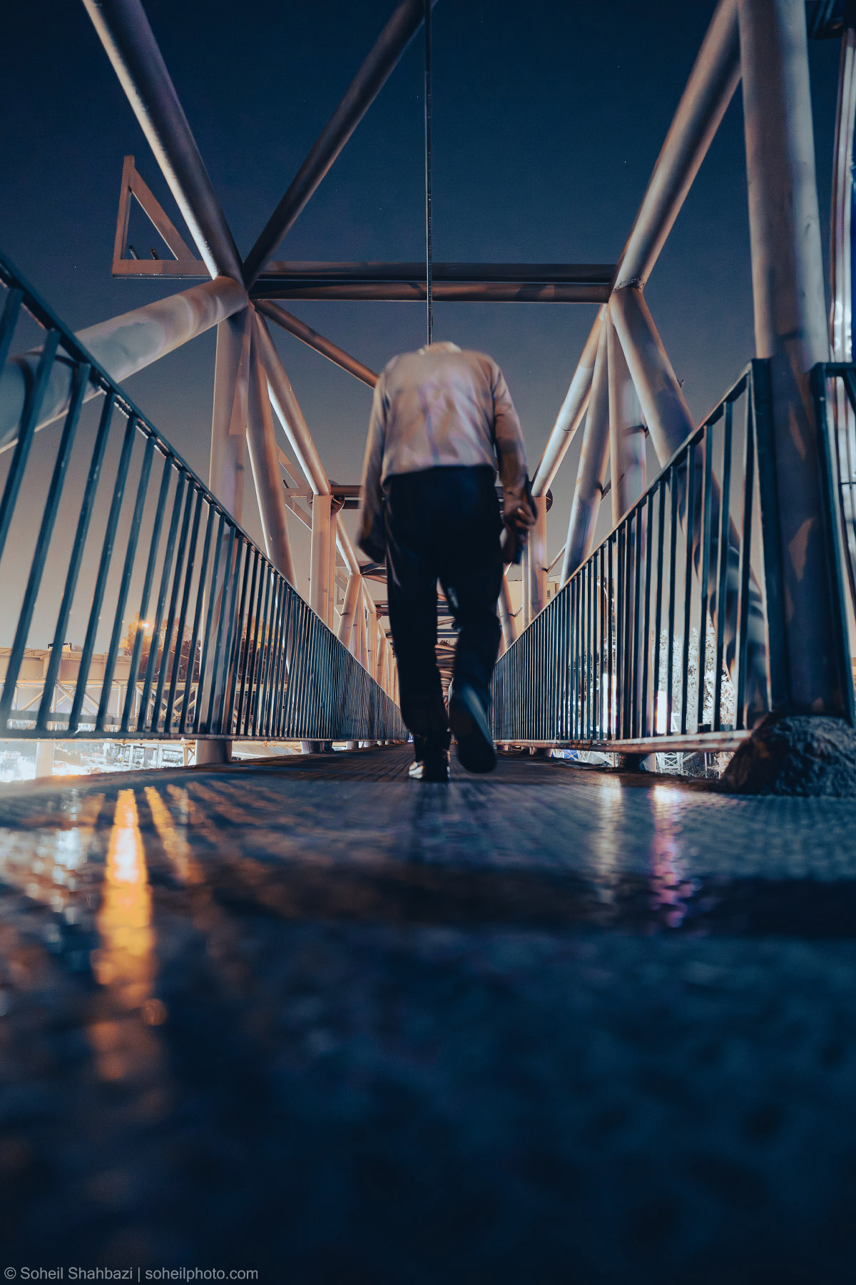 A low-angle, cinematic shot of a person walking away on a modern pedestrian bridge with geometric steel beams, captured with deep blue tones and reflections on the wet ground. Photographed by Soheil Shahbazi