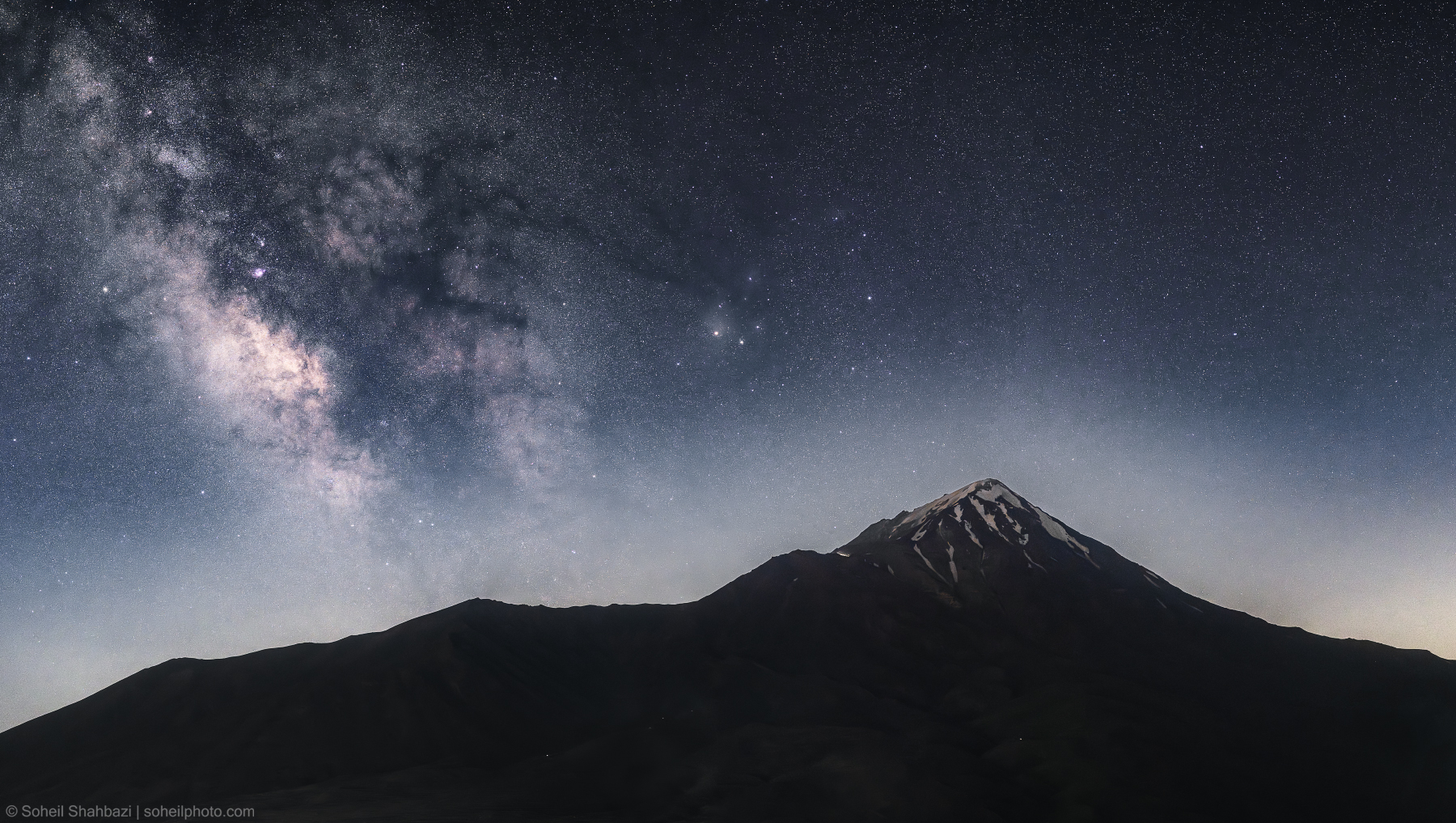A breathtaking wide-angle night landscape of Mount Damavand peak under a vivid Milky Way galaxy sky, showcasing the starry cosmos and the snowy summit of the highest peak in Iran. Photographed by Soheil Shahbazi