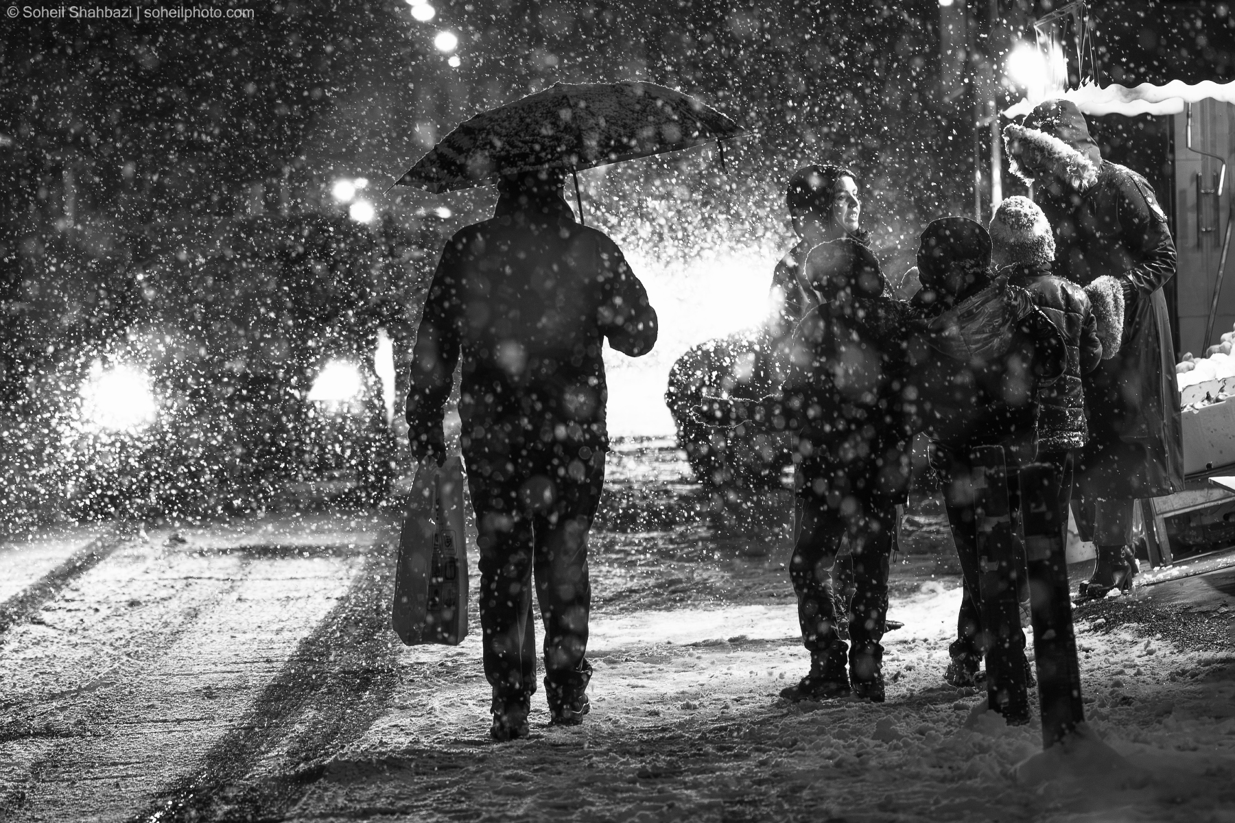 Alt text: A dramatic black and white street scene during a heavy snowfall, featuring a person holding an umbrella and a shopping bag, standing near a group of people in a snowy urban environment. Photographed by Soheil Shahbazi