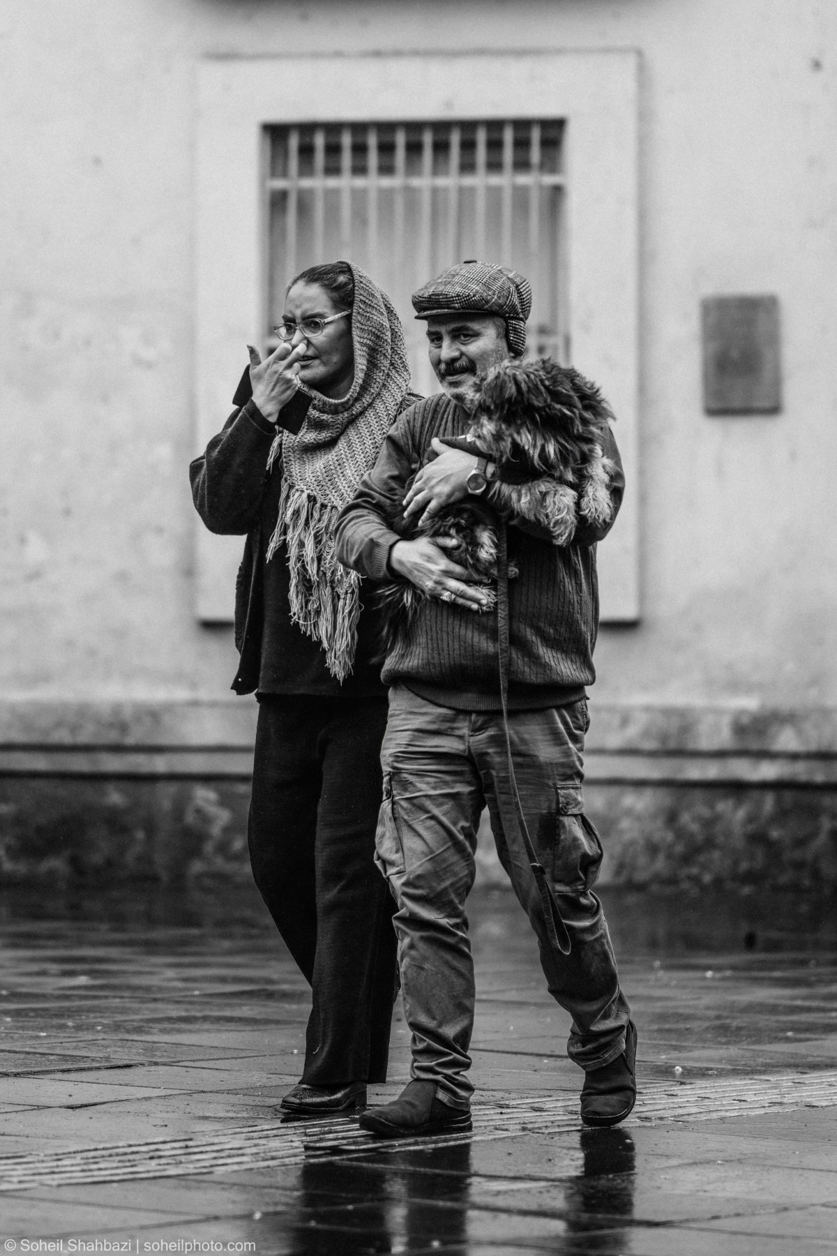 A candid black and white street portrait of an older couple walking together, with the man wearing a flat cap and holding a small fluffy dog in his arms. Photographed by Soheil Shahbazi