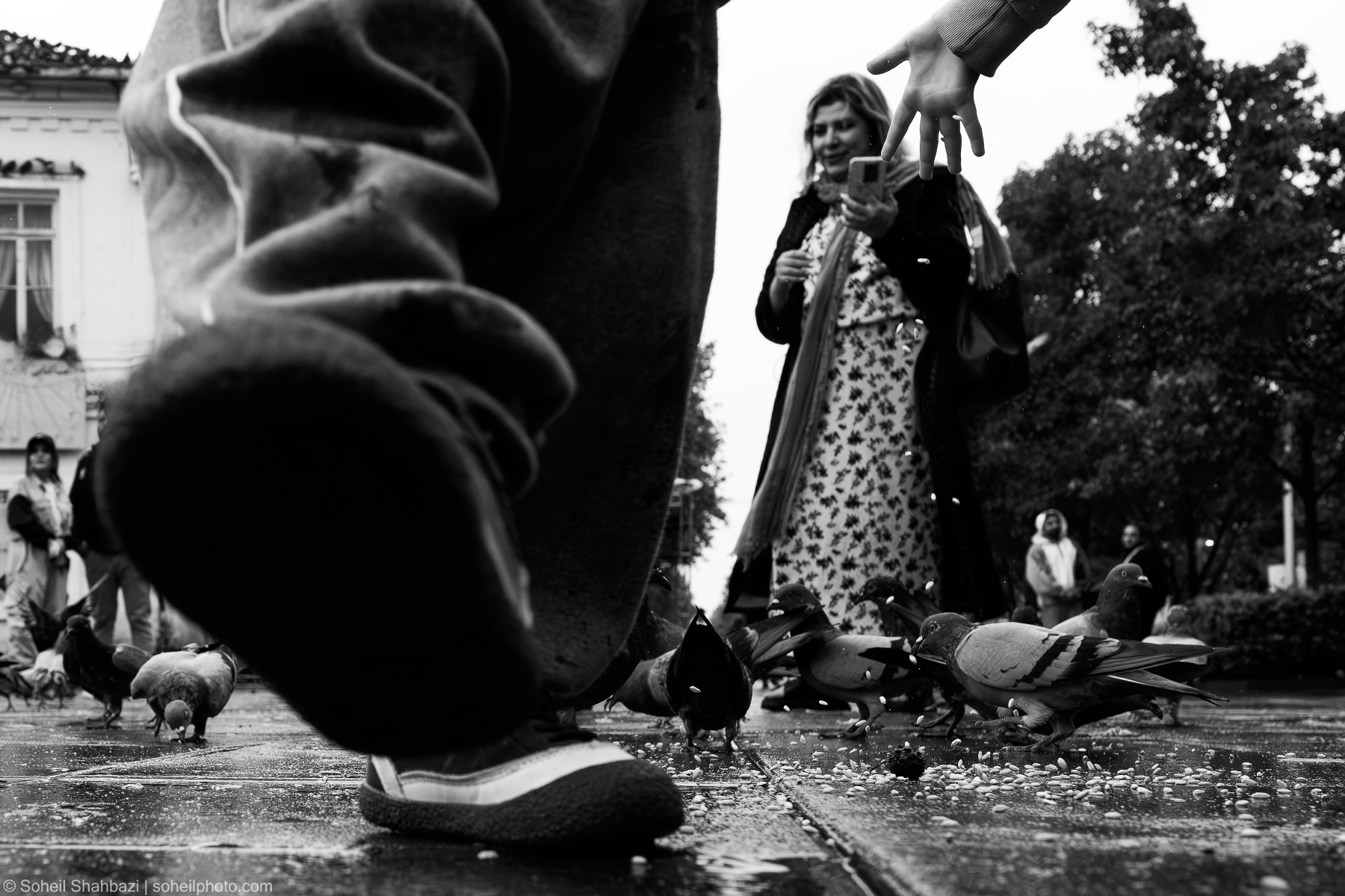 A low-angle, black and white street photography shot showing a person's foot in the foreground, with a woman feeding pigeons in a public square in the background. Photographed by Soheil Shahbazi